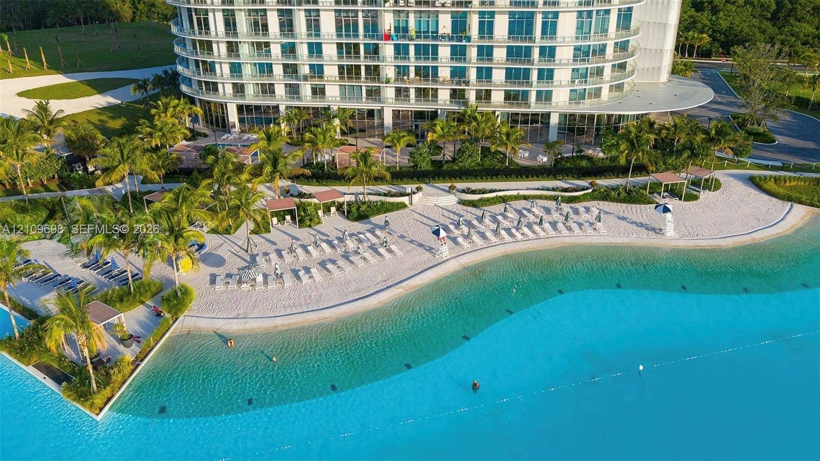 2411 Laguna Circle, #810, North Miami, FL 33181 Aerial perspective of high-rise structure featuring a crystalline swimming lagoon, expansive sand beach, paved pathways, mature palm landscaping, and dedicated shaded cabanas