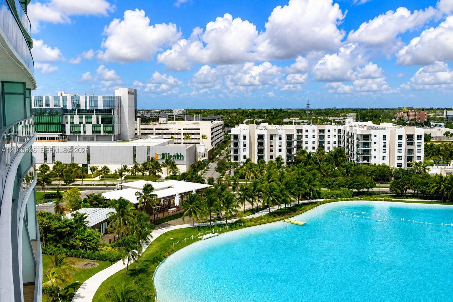 2411 Laguna Circle, #810, North Miami, FL 33181 Elevated balcony featuring a glass railing, overlooking a large turquoise lagoon, landscaped grounds, palm trees, and paved walking paths