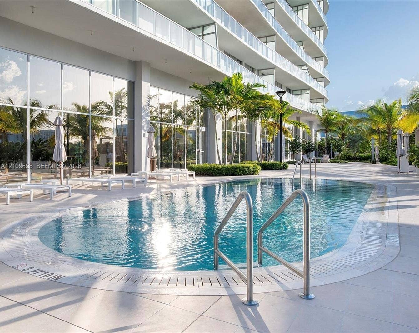 2411 Laguna Circle, #810, North Miami, FL 33181 Swimming pool with metal handrails, expansive tile pool deck, floor-to-ceiling glass wall facade, and tropical landscaping