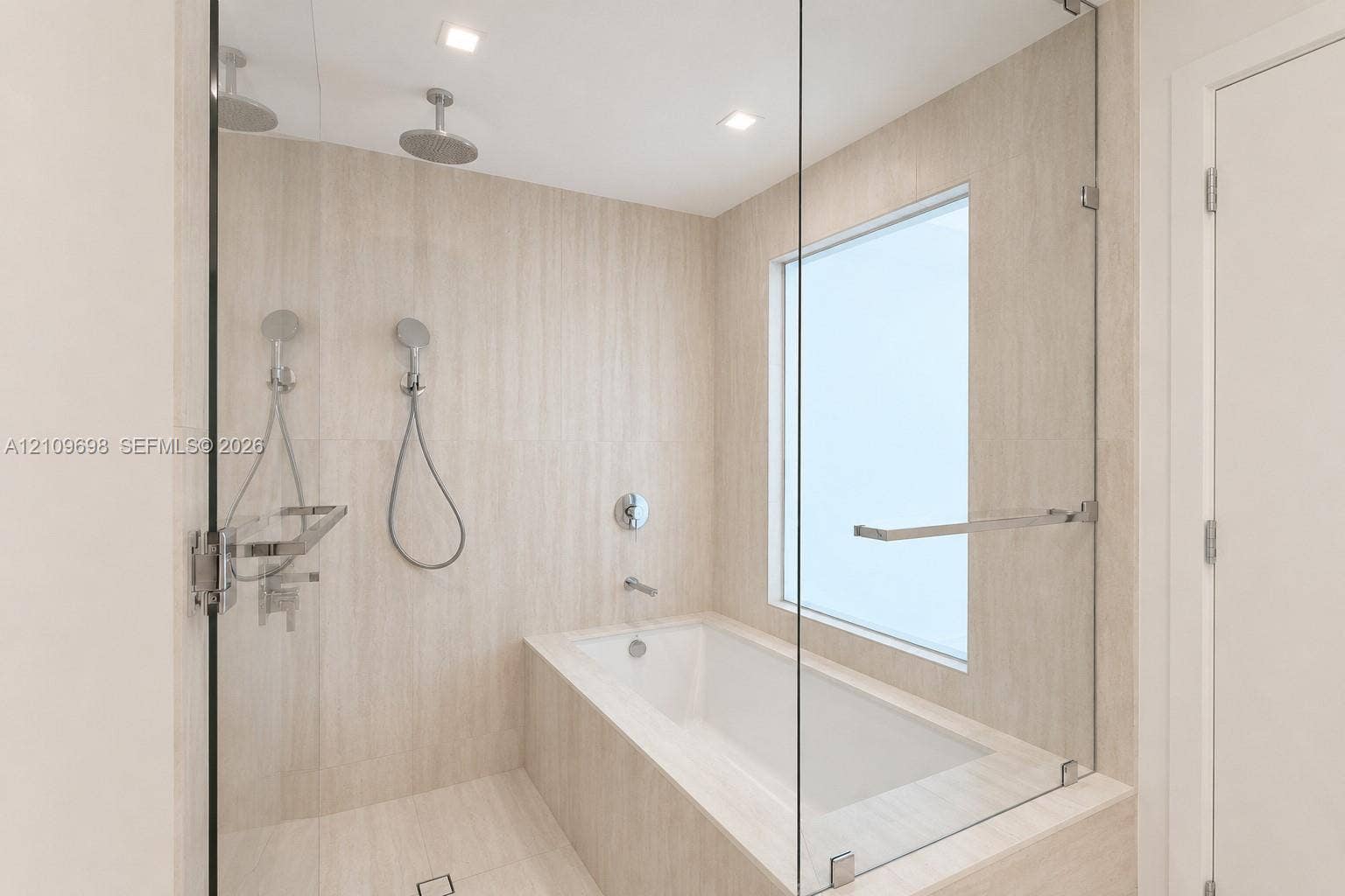 2411 Laguna Circle, #810, North Miami, FL 33181 Enclosed wet room featuring a deep soaking tub, frameless glass partition, dual wall-mounted rainfall showerheads, and neutral-toned stone-finish tiling