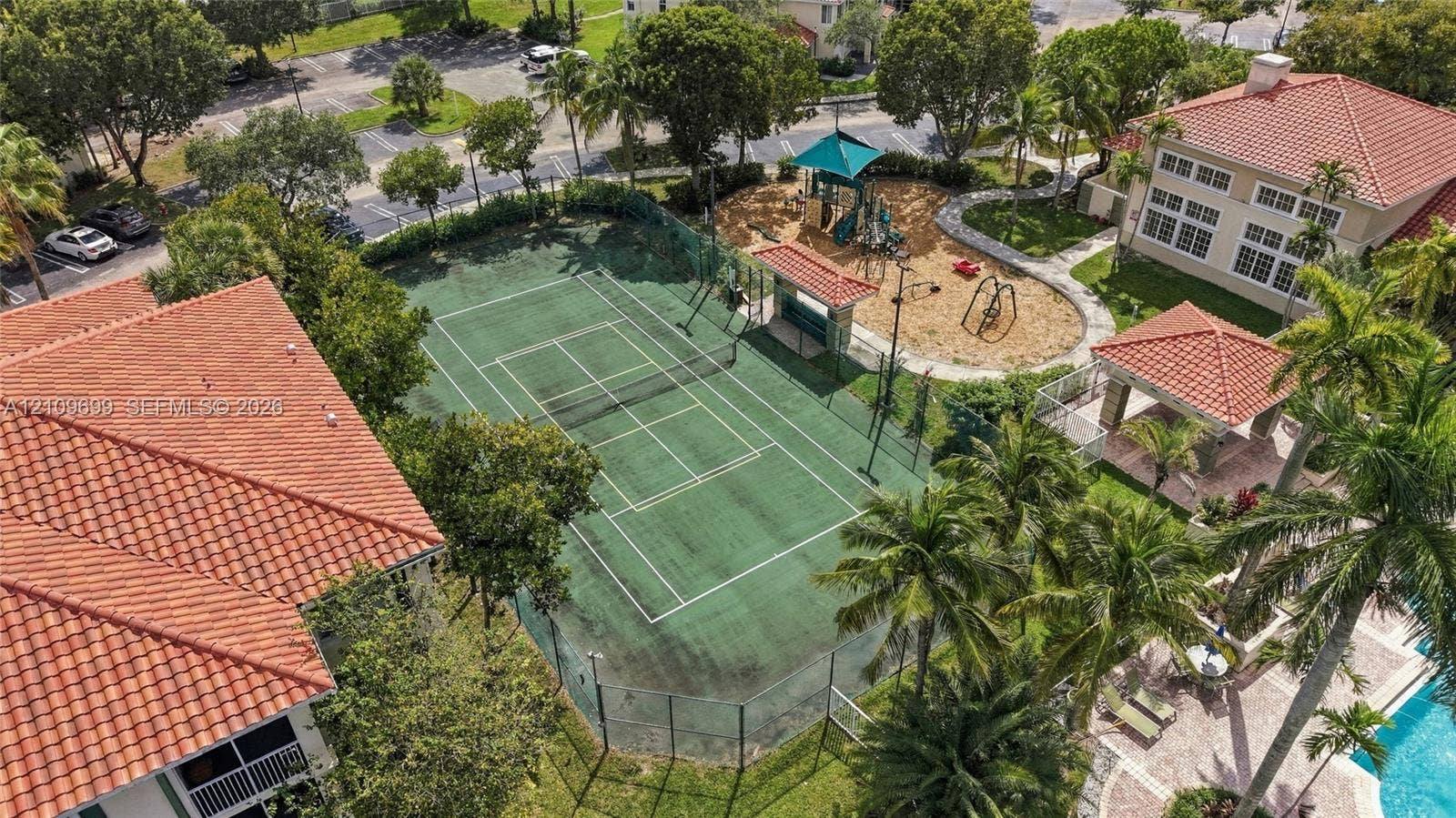 Tennis court with perimeter fencing, outdoor play area with structures, covered pavilion, and surrounding palm trees