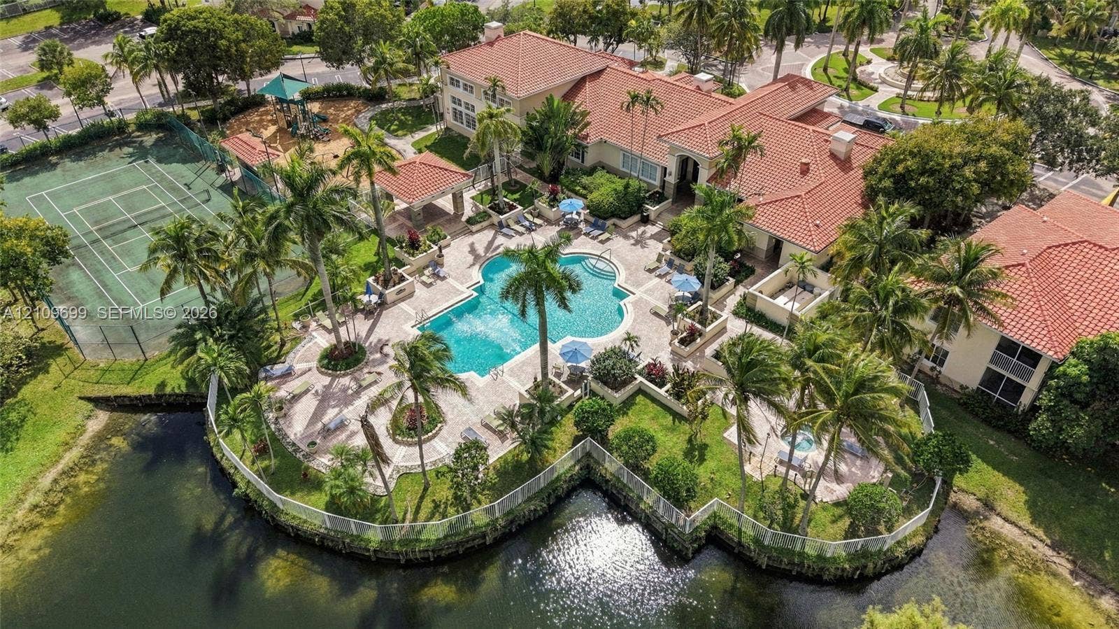 Aerial perspective of a sprawling estate featuring a terracotta-tiled roof, a custom-shaped swimming pool, a private tennis court, a fenced waterfront perimeter, and integrated spa