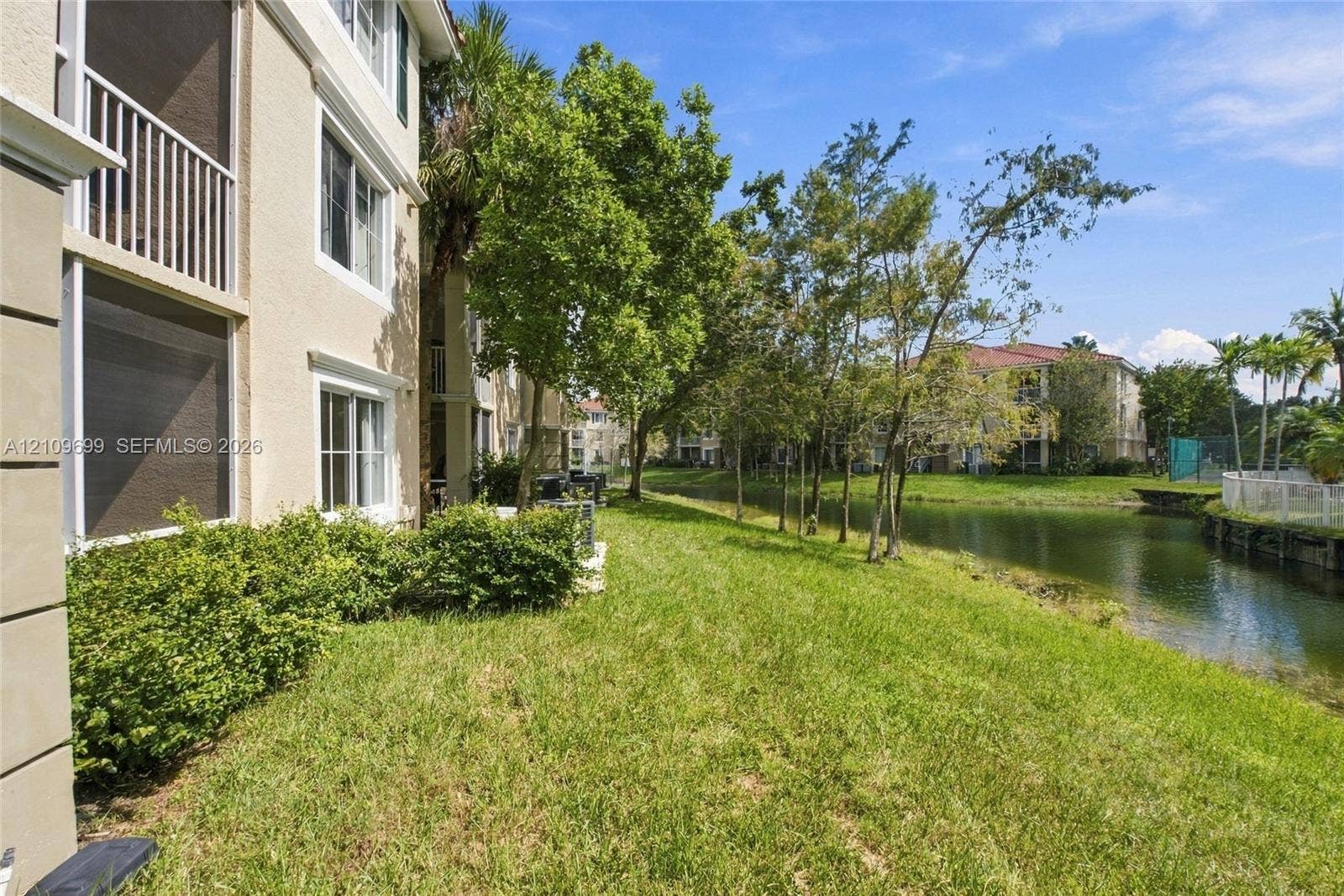 Multi-story stucco exterior featuring screened balconies, landscaped lawn, mature trees, and water frontage