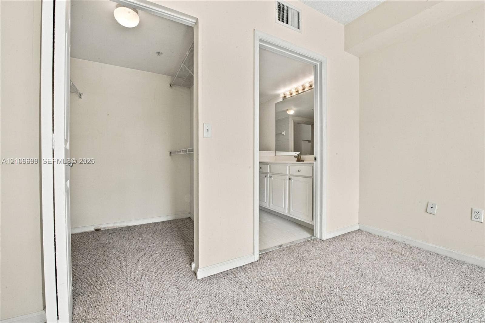 Walk-in closet featuring wire shelving and overhead lighting, with an entry to a bathroom equipped with white cabinetry, a large vanity mirror, and multi-bulb wall lighting