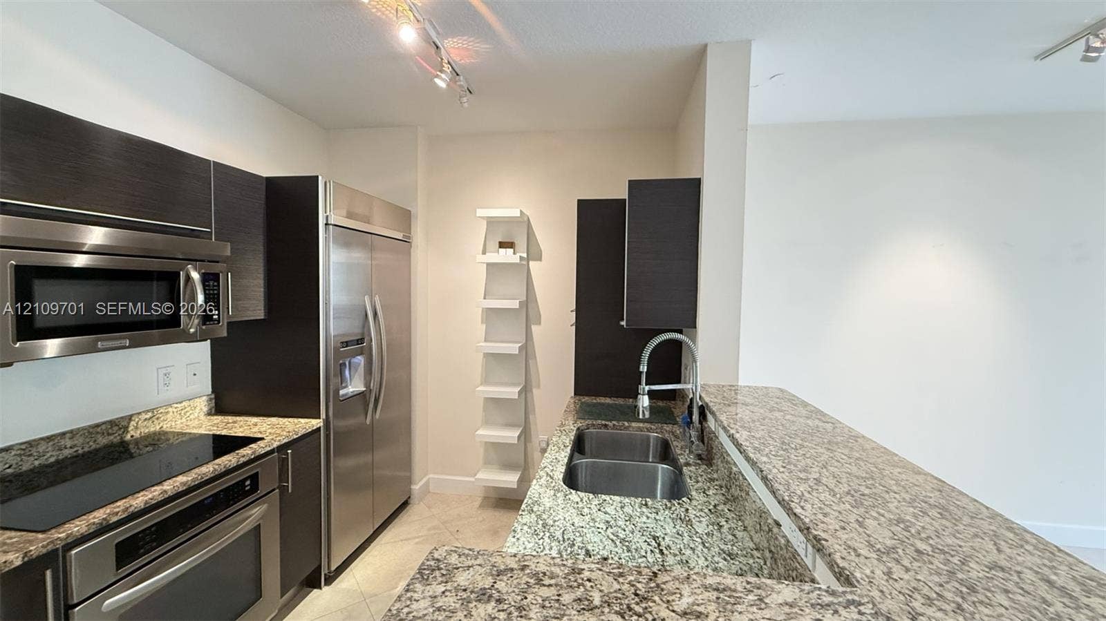 Kitchen featuring dark cabinetry, stone-finish countertops, stainless steel appliances, a double-basin undermount sink, and built-in vertical shelving