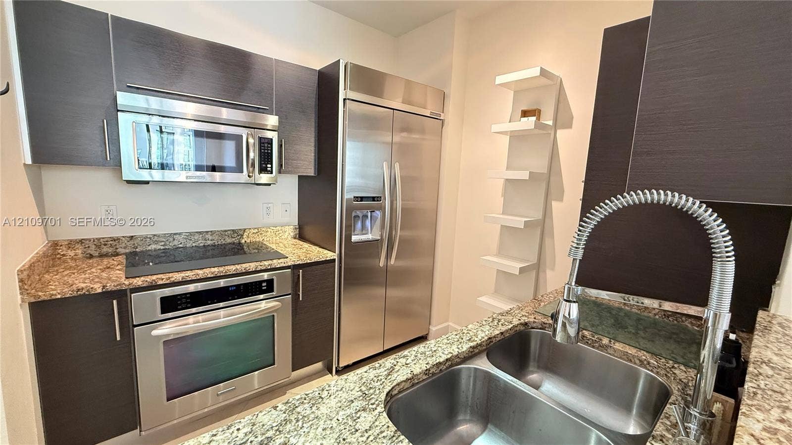 Kitchen featuring dark wood-finish cabinetry, speckled stone countertops, a stainless steel side-by-side refrigerator, a built-in microwave, and a double-basin stainless steel sink with a high-arch spring faucet