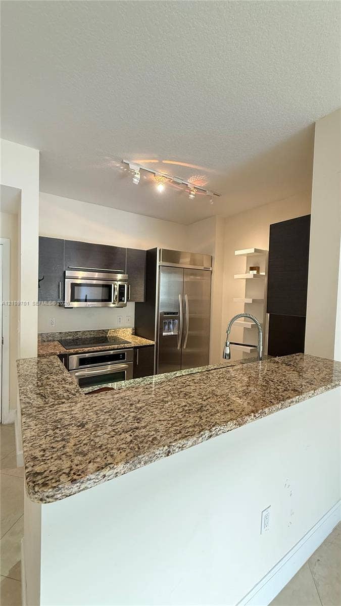 Kitchen featuring stone countertops, stainless steel appliances, dark cabinetry, track lighting, and built-in shelving