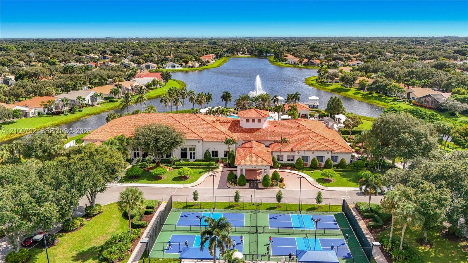 Aerial perspective of a multi-court pickleball complex with perimeter fencing, stadium-style lighting, and paved pathways, situated on a lakefront lot with a tiled-roof clubhouse and a central fountain