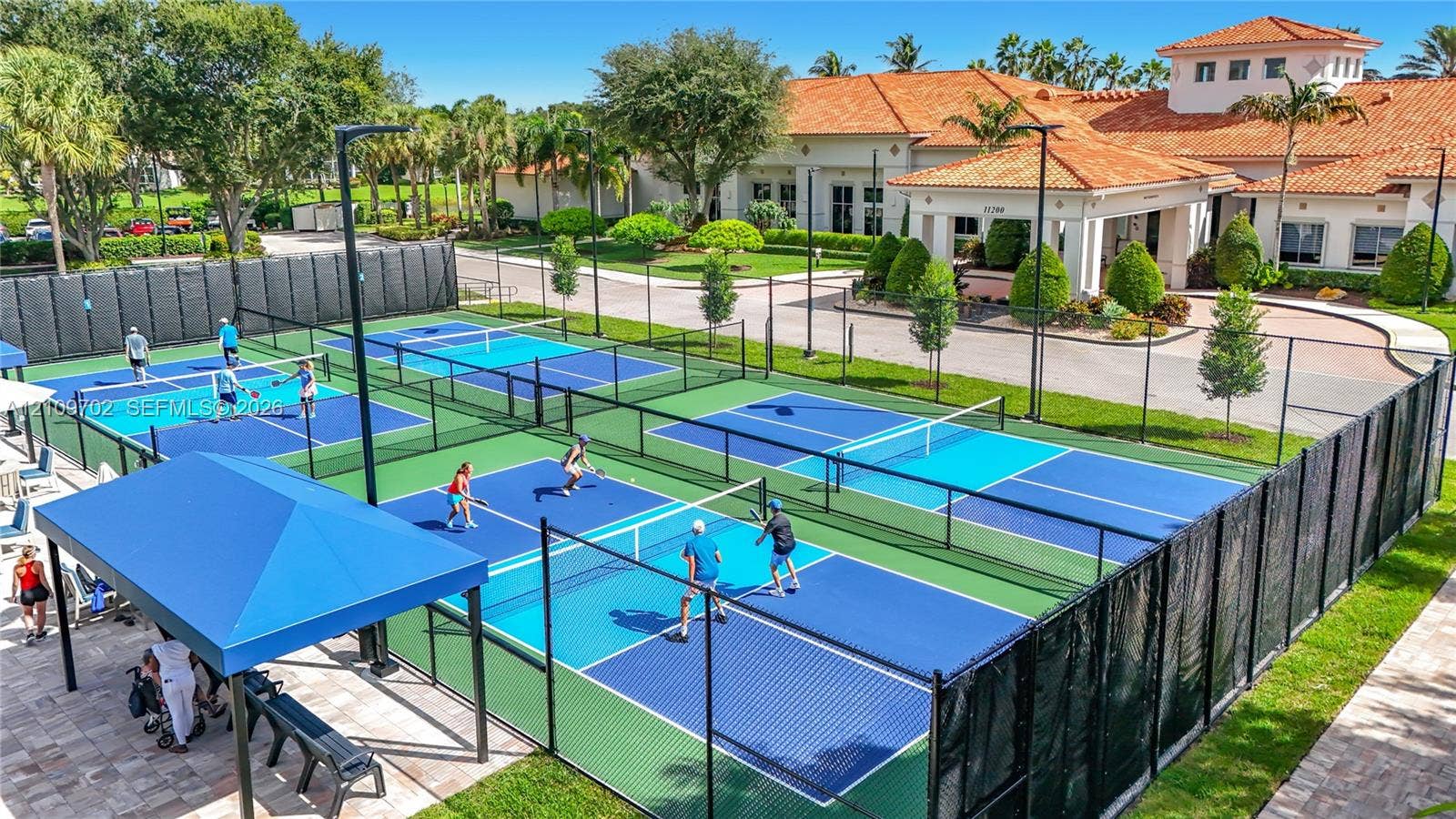 Multi-court pickleball facility featuring high-perimeter fencing, integrated court lighting, a covered seating area with shade canopy, and paver-stone pathways