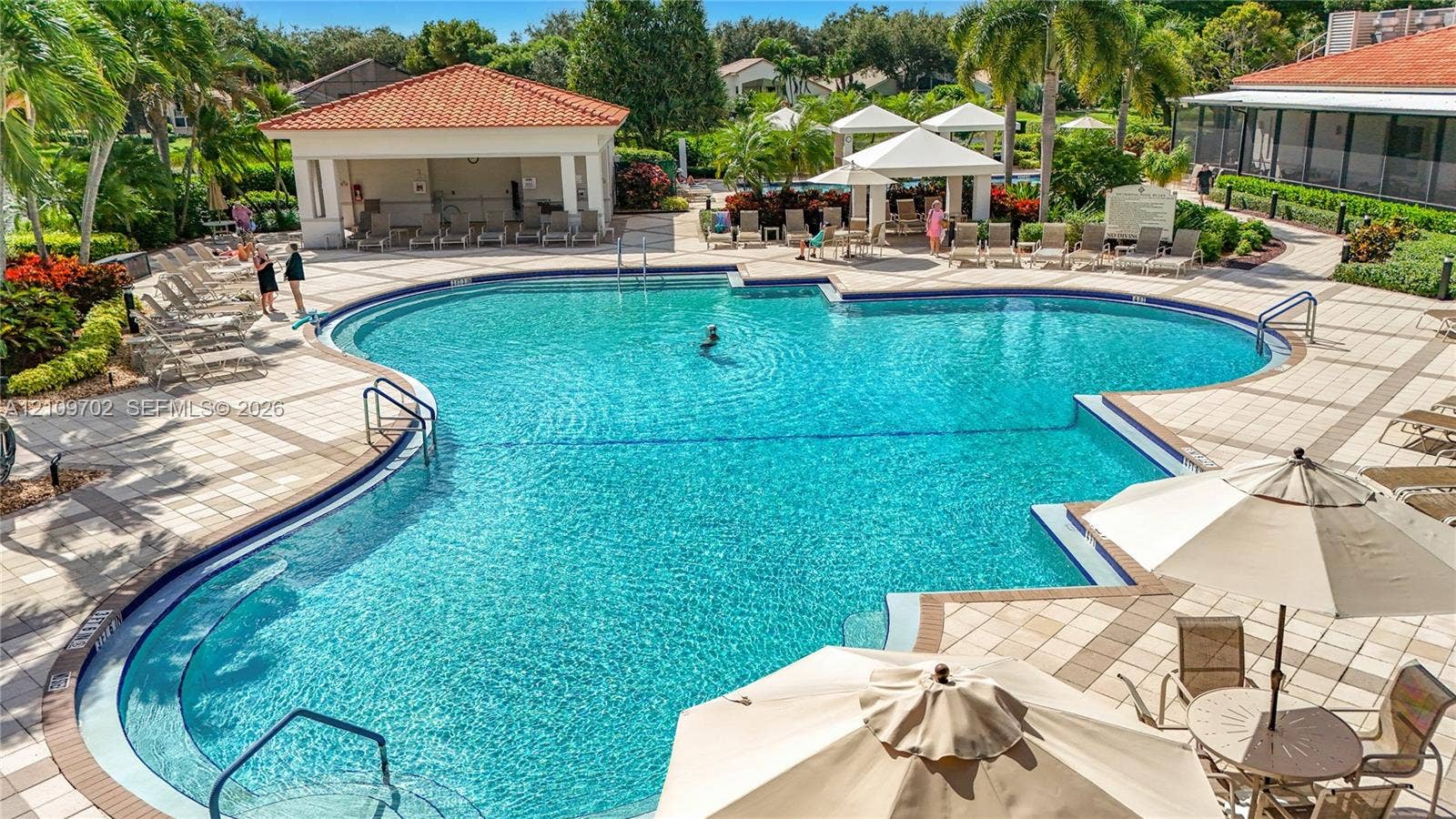 Community swimming pool featuring a geometric design, expansive paved pool deck, and shaded pavilion structures with terracotta-style roof tiles