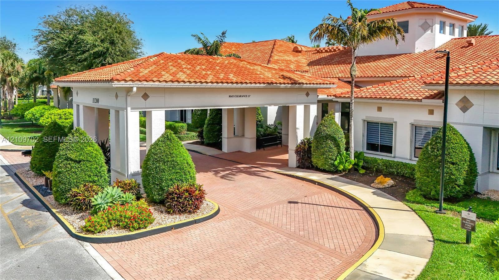 Mediterranean-style exterior featuring a covered porte-cochere, clay tile roofing, structured landscaping, and a brick-paver circular driveway