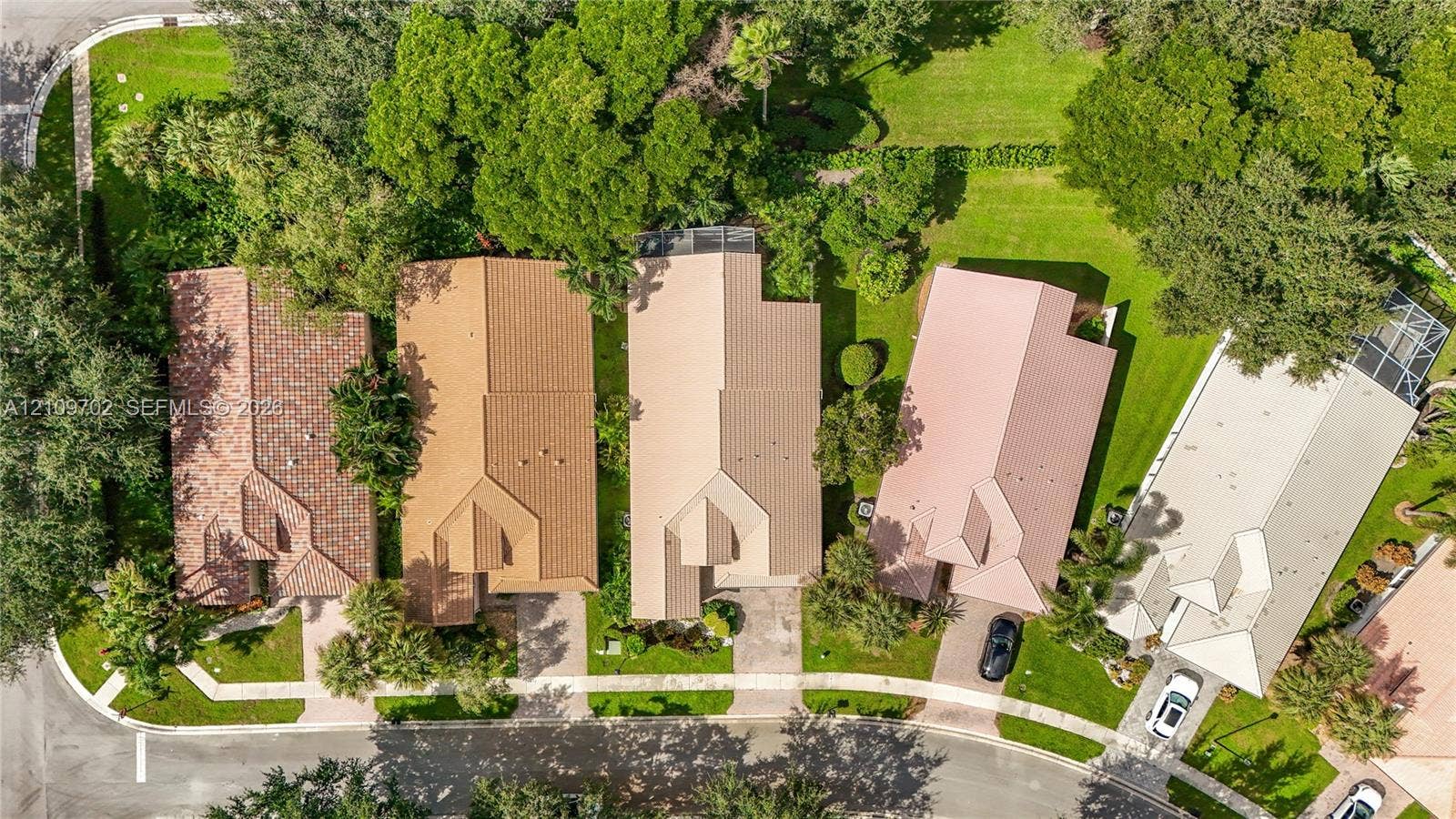 Aerial view featuring mature tree canopy, manicured lawns, paved driveways, and tile roof structures