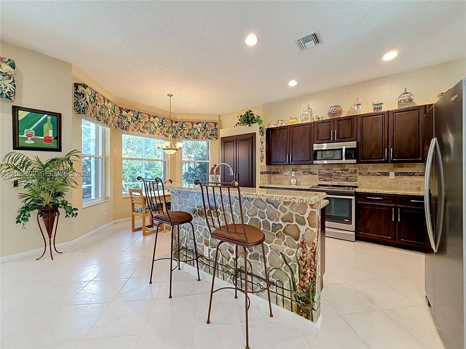 Kitchen featuring dark wood cabinetry, stone-veneer breakfast bar with integrated sink, stone tile backsplash, stainless steel appliances, and large windows with integrated dining area