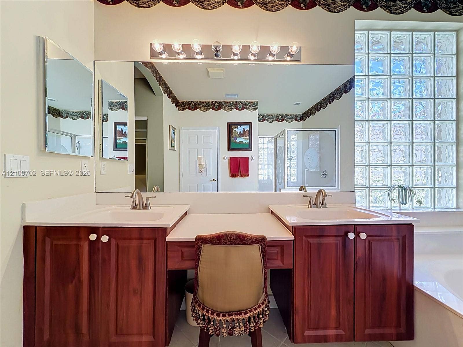 Dual-sink vanity with wood-finish cabinetry, solid surface countertop, glass block window, and large wall-to-wall mirror
