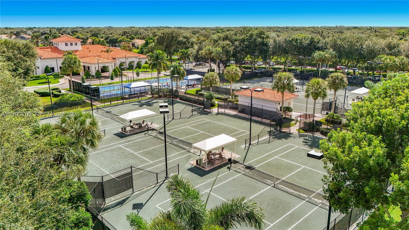 Aerial view of multiple fenced tennis courts featuring hard-court surfaces, elevated lighting, and shaded seating structures