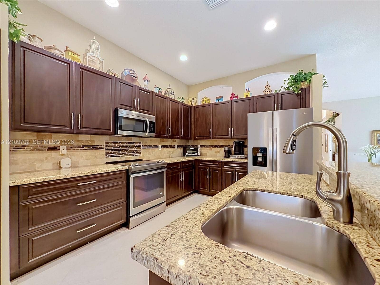 Kitchen featuring dark wood cabinetry, light stone countertops, stainless steel appliances, a double-basin undermount sink with a gooseneck faucet, and a stone tile backsplash