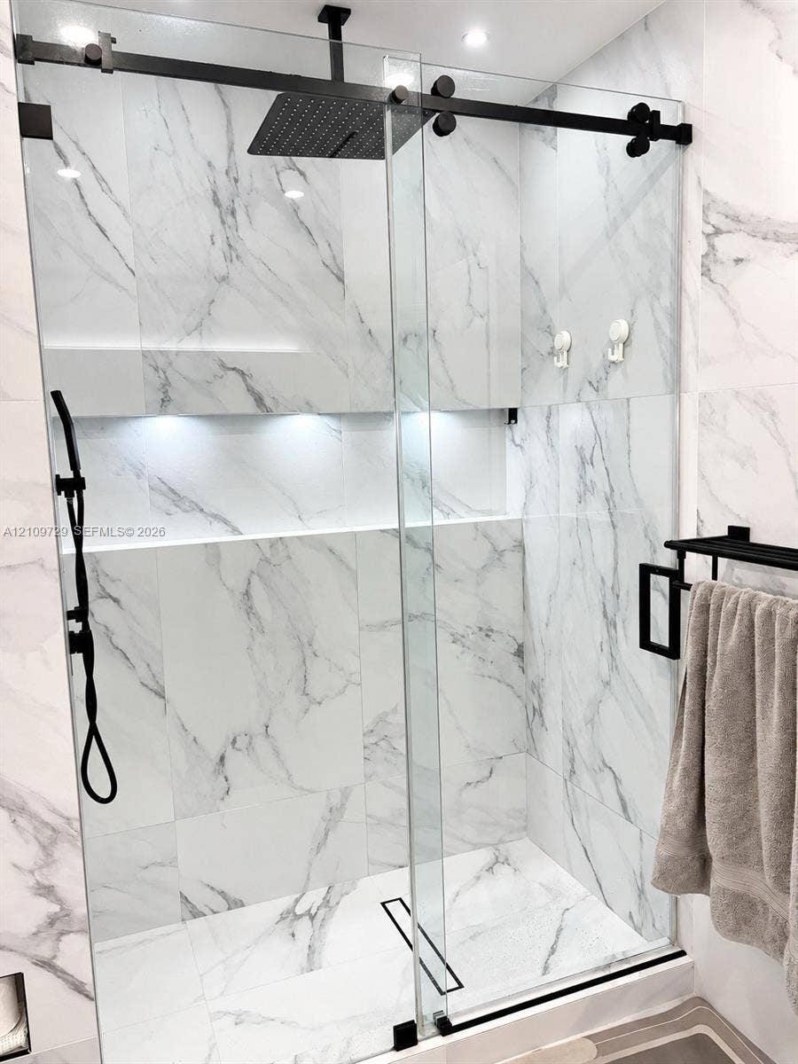 12665 SW 54th Court, Miramar, FL 33027 Walk-in shower featuring large-format stone-finish wall and floor tile, glass sliding door enclosure, black rainfall shower head with handheld attachment, and an integrated recessed shelf