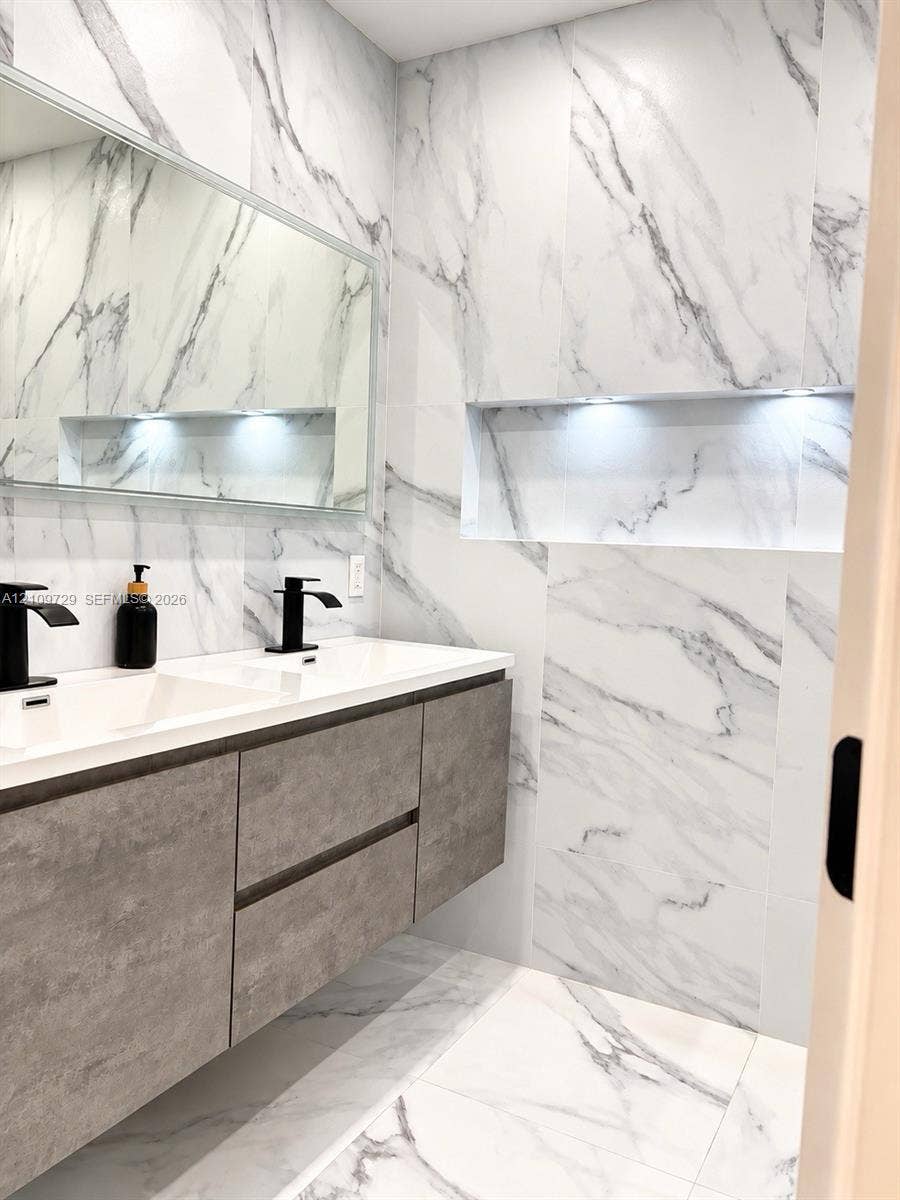 12665 SW 54th Court, Miramar, FL 33027 Floating vanity with gray-textured cabinetry, double-sink countertop, matte black fixtures, full-wall stone-patterned tile, and recessed wall niche with integrated lighting