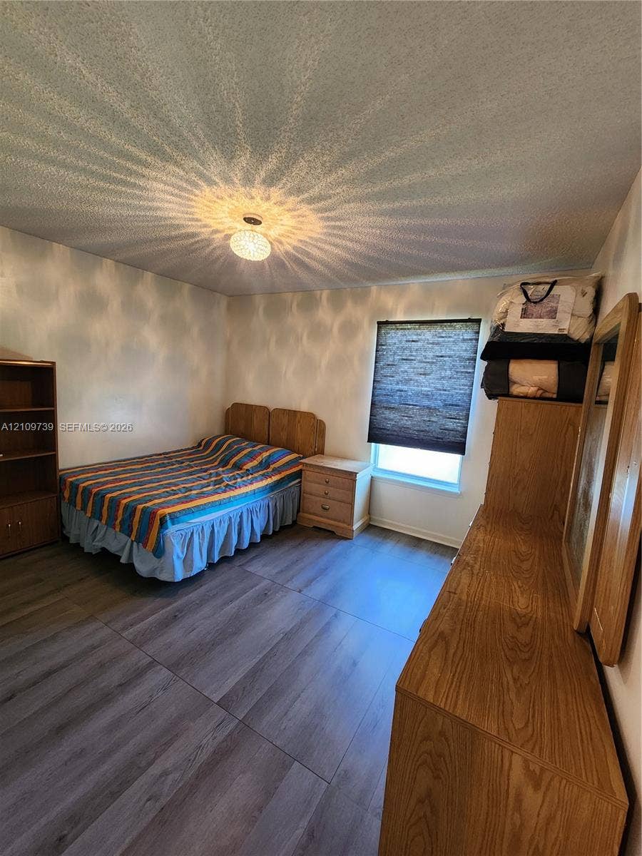Wood-finish flooring, textured ceiling, central ceiling-mounted light fixture, and window with adjustable shade