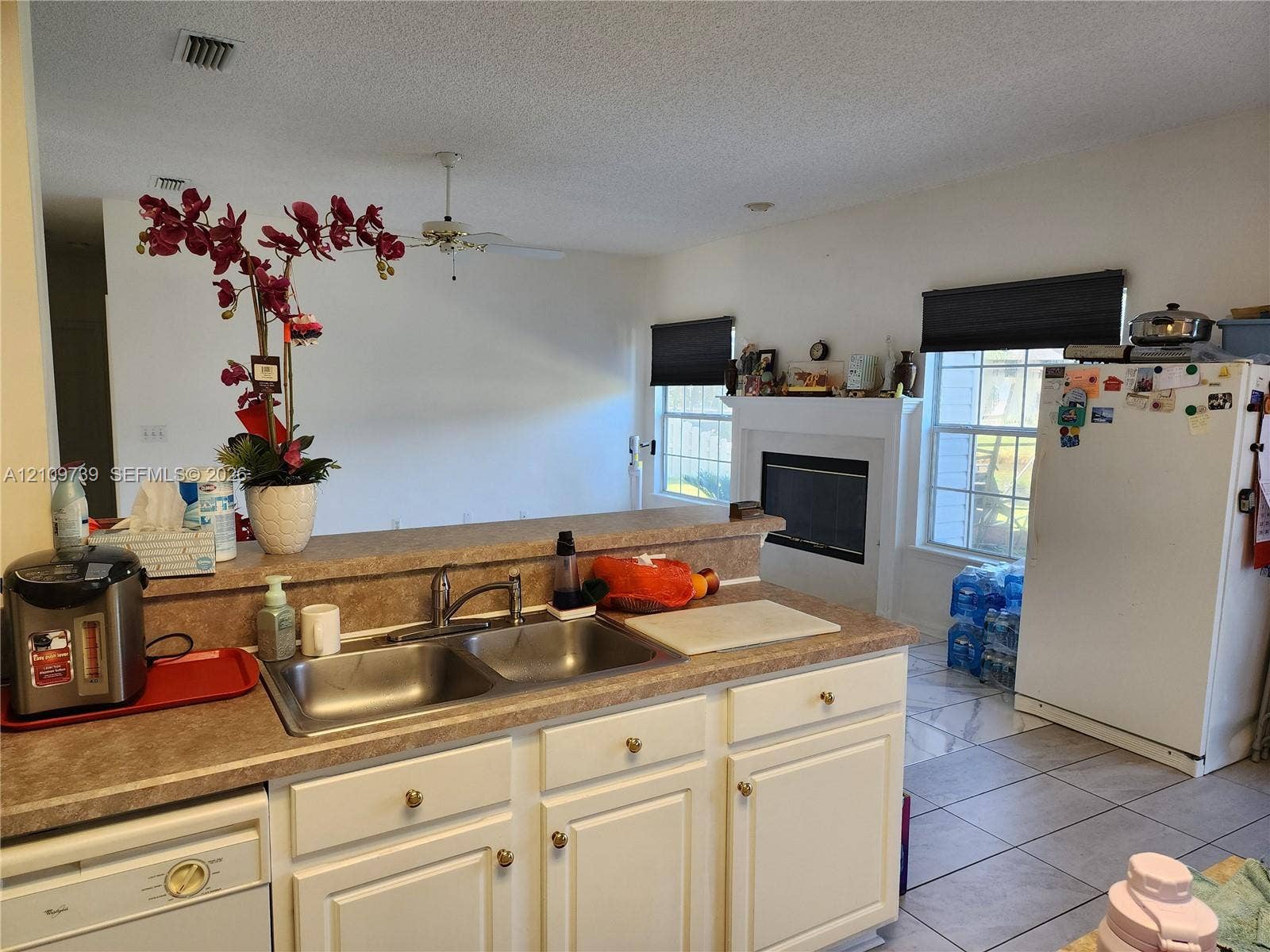 Kitchen featuring white cabinetry, solid-surface countertops, dual-basin stainless steel sink, tile flooring, and a fireplace with a white mantel