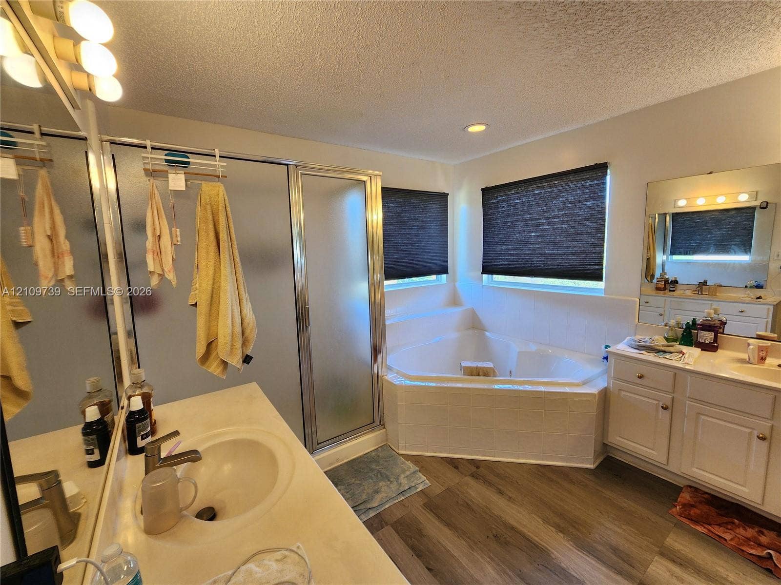 Bathroom featuring wood-finish flooring, a corner soaking tub with tiled surround, a separate glass-enclosed shower, dual vanity cabinets, and a framed mirror