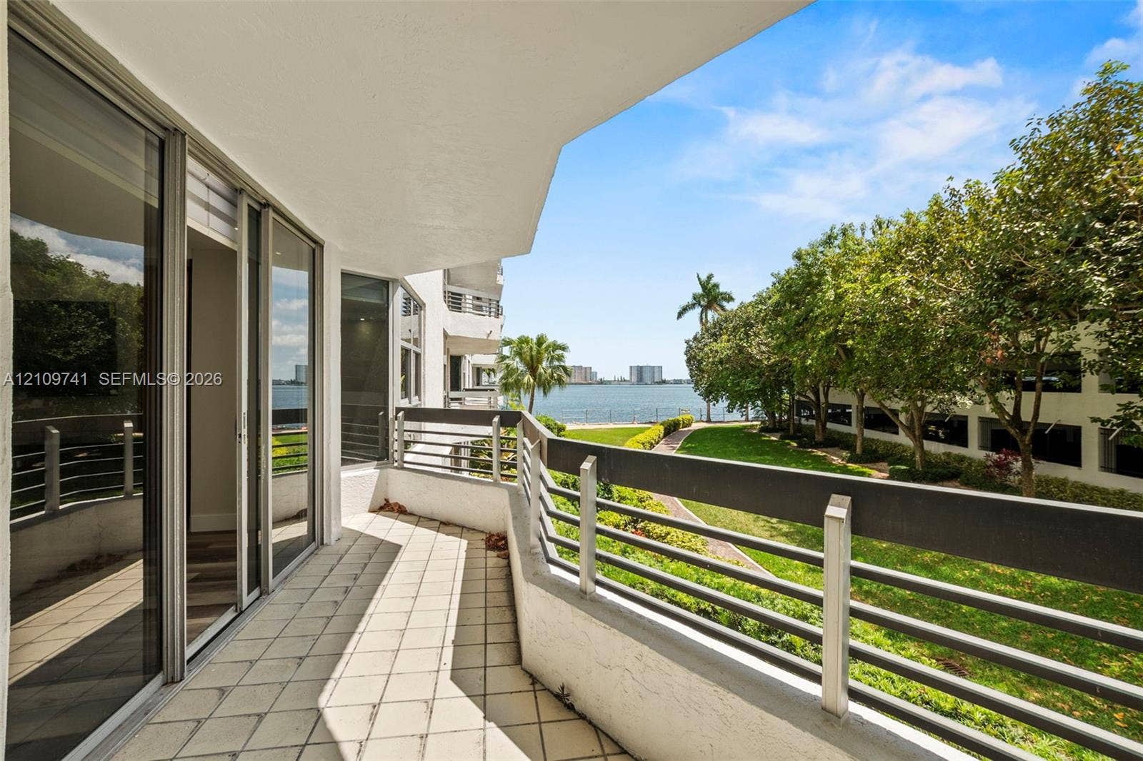 19101 NE 36th Court, #203, Aventura, FL 33180 Covered balcony featuring tile flooring, metal railings, and sliding glass door entry, with water frontage and mature landscaping