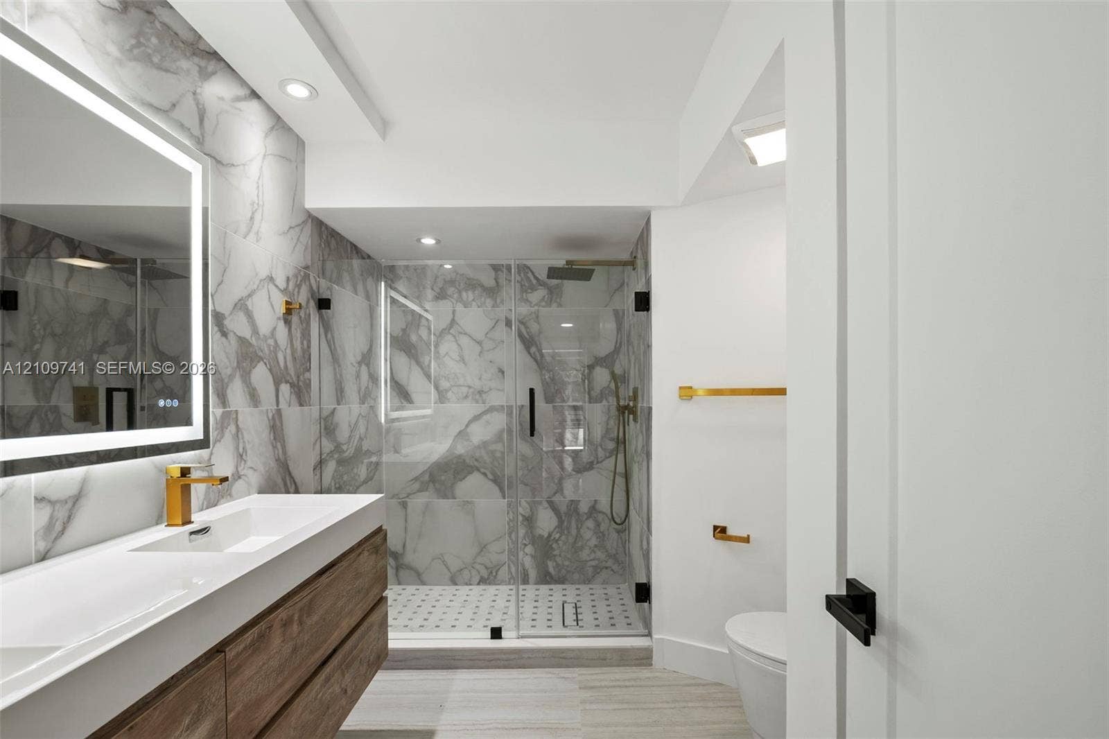 19101 NE 36th Court, #203, Aventura, FL 33180 Floating wood-finish vanity with an integrated sink, backlit LED mirror, walk-in shower with stone-patterned tile, and gold-tone fixtures