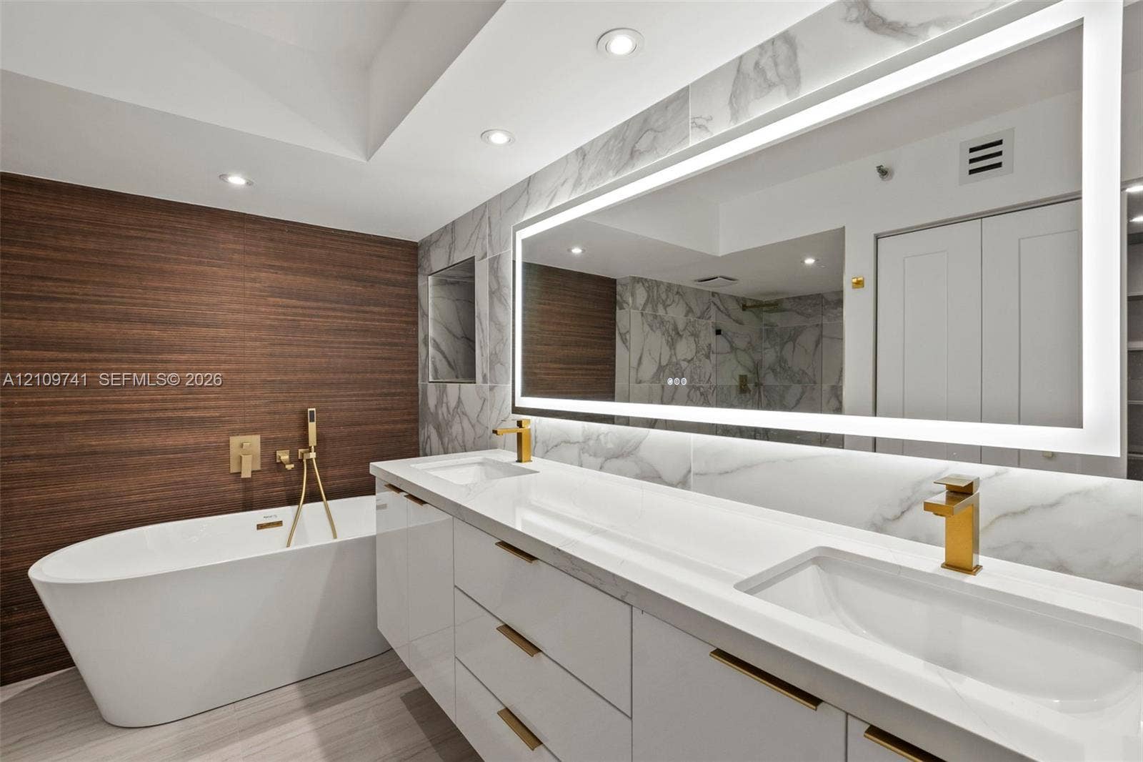 19101 NE 36th Court, #203, Aventura, FL 33180 Freestanding soaking tub with wall-mounted gold-toned hardware, white vanity with integrated double sinks, large LED-backlit mirror, wood-finish feature wall, and stone-finish wall tile