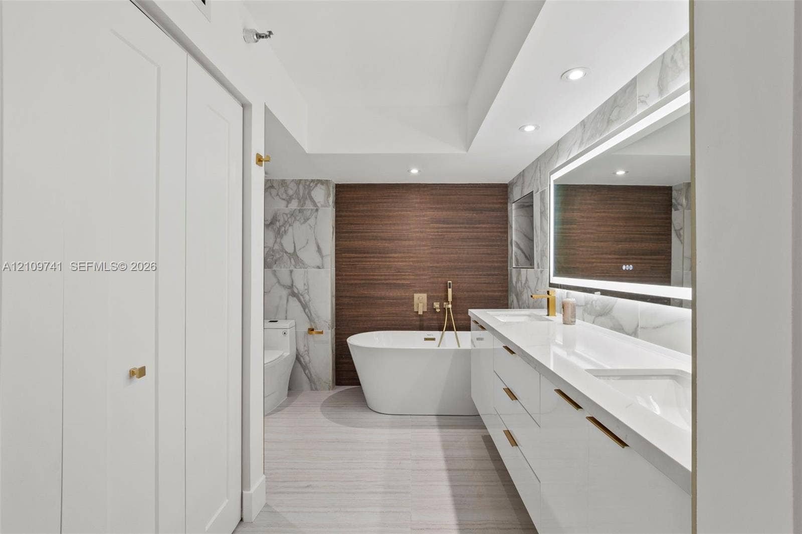 19101 NE 36th Court, #203, Aventura, FL 33180 Modern bathroom featuring a dual-sink vanity with white cabinetry, freestanding soaking tub with wall-mounted gold-tone hardware, large-format tiled walls, and LED-backlit mirror