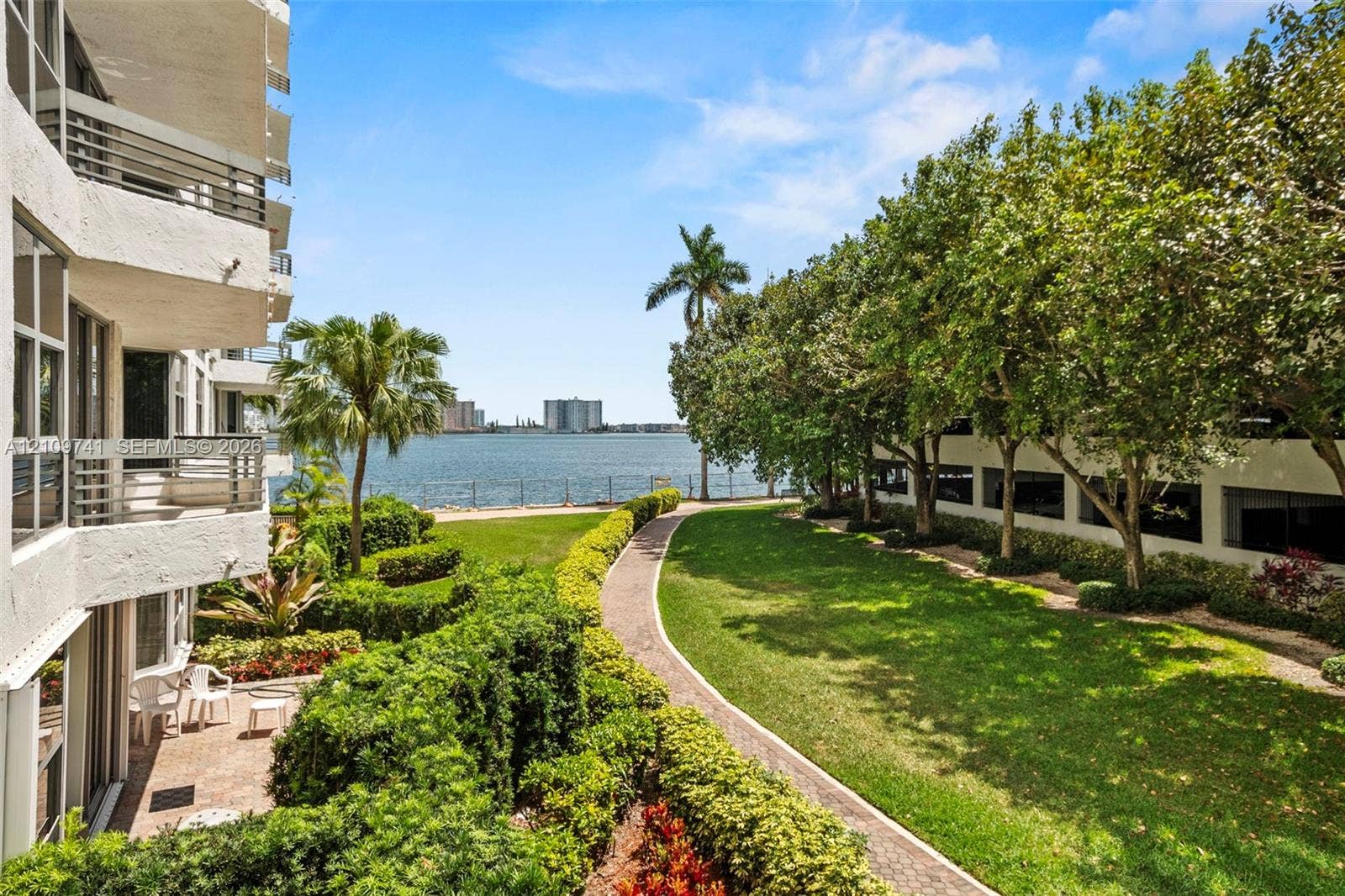 19101 NE 36th Court, #203, Aventura, FL 33180 Waterfront grounds featuring a winding stone-paver path, manicured lawn, mature trees, and private balconies with horizontal railings