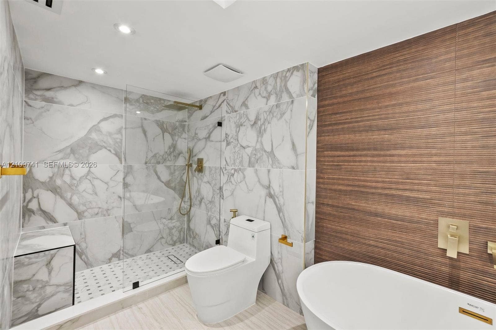 19101 NE 36th Court, #203, Aventura, FL 33180 Full-height stone-patterned tile shower enclosure with integrated bench and frameless glass door, freestanding soaking tub, wood-finish textured accent wall, and gold-toned wall-mounted plumbing fixtures