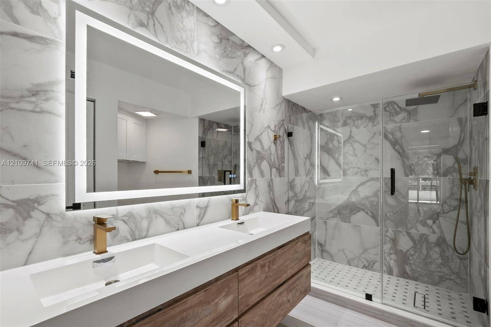 19101 NE 36th Court, #203, Aventura, FL 33180 Floating dual-sink vanity with wood-finish cabinetry, backlit LED mirror, large-format stone-patterned tile wall, and glass-enclosed shower with gold-tone hardware