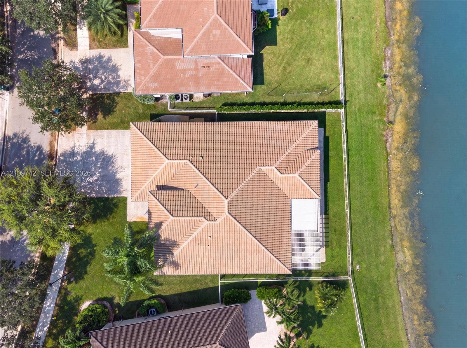 19143 SW 60th Court, Pembroke Pines, FL 33332 Aerial perspective of a residential property featuring a multi-hipped tile roof, concrete driveway, and expansive lawn area with perimeter fencing along the waterfront