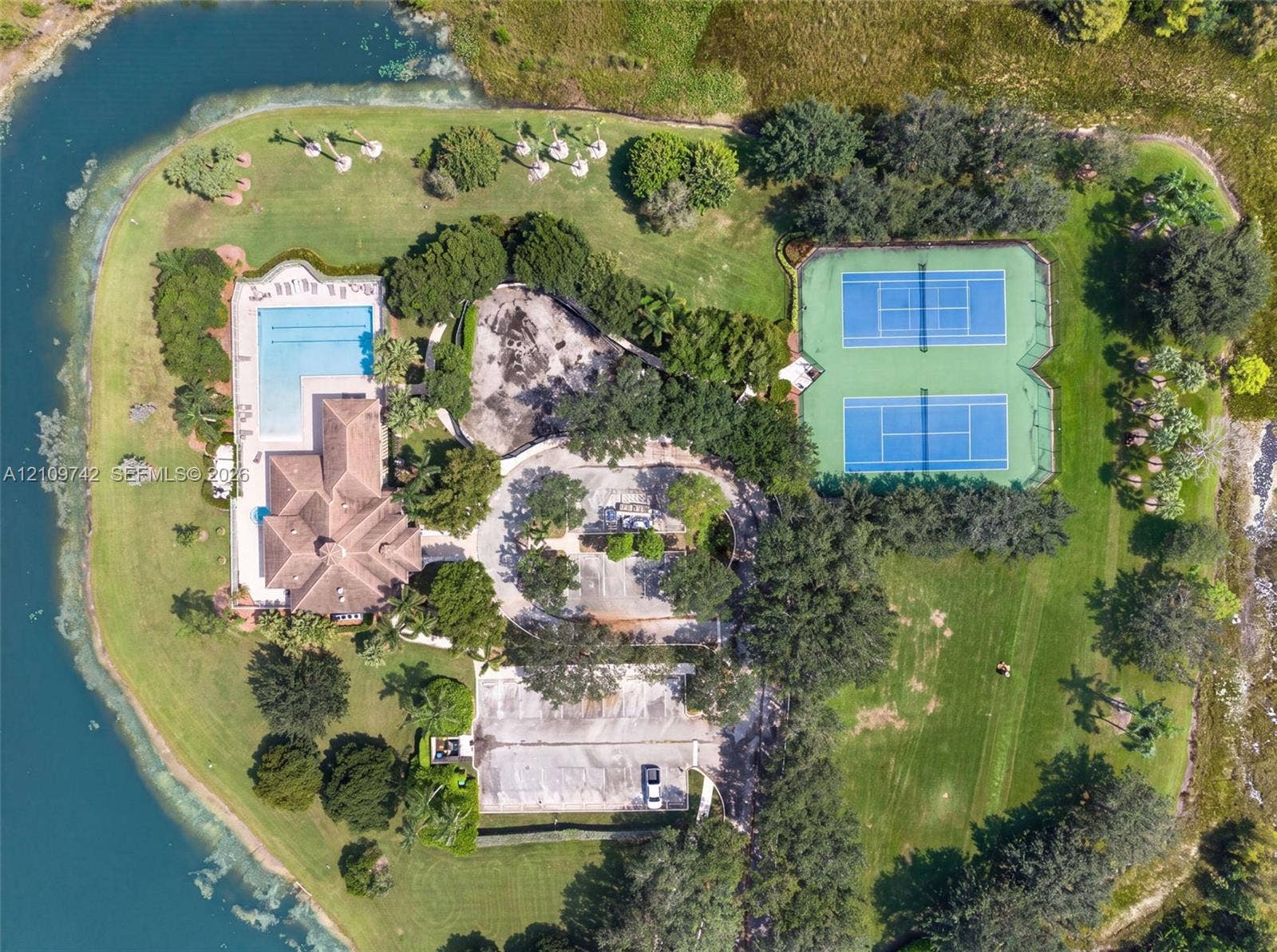 19143 SW 60th Court, Pembroke Pines, FL 33332 Aerial perspective of a multi-acre property featuring a swimming pool, two hard-court tennis courts, expansive manicured lawn, and water frontage