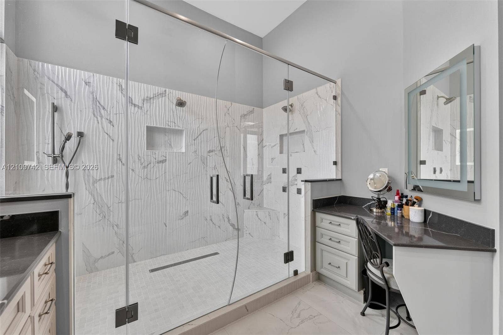 19143 SW 60th Court, Pembroke Pines, FL 33332 Walk-in frameless glass shower featuring textured tile surround, built-in bench, dual shower heads, and multiple body sprays, complemented by dual vanities with dark stone countertops and an illuminated mirror