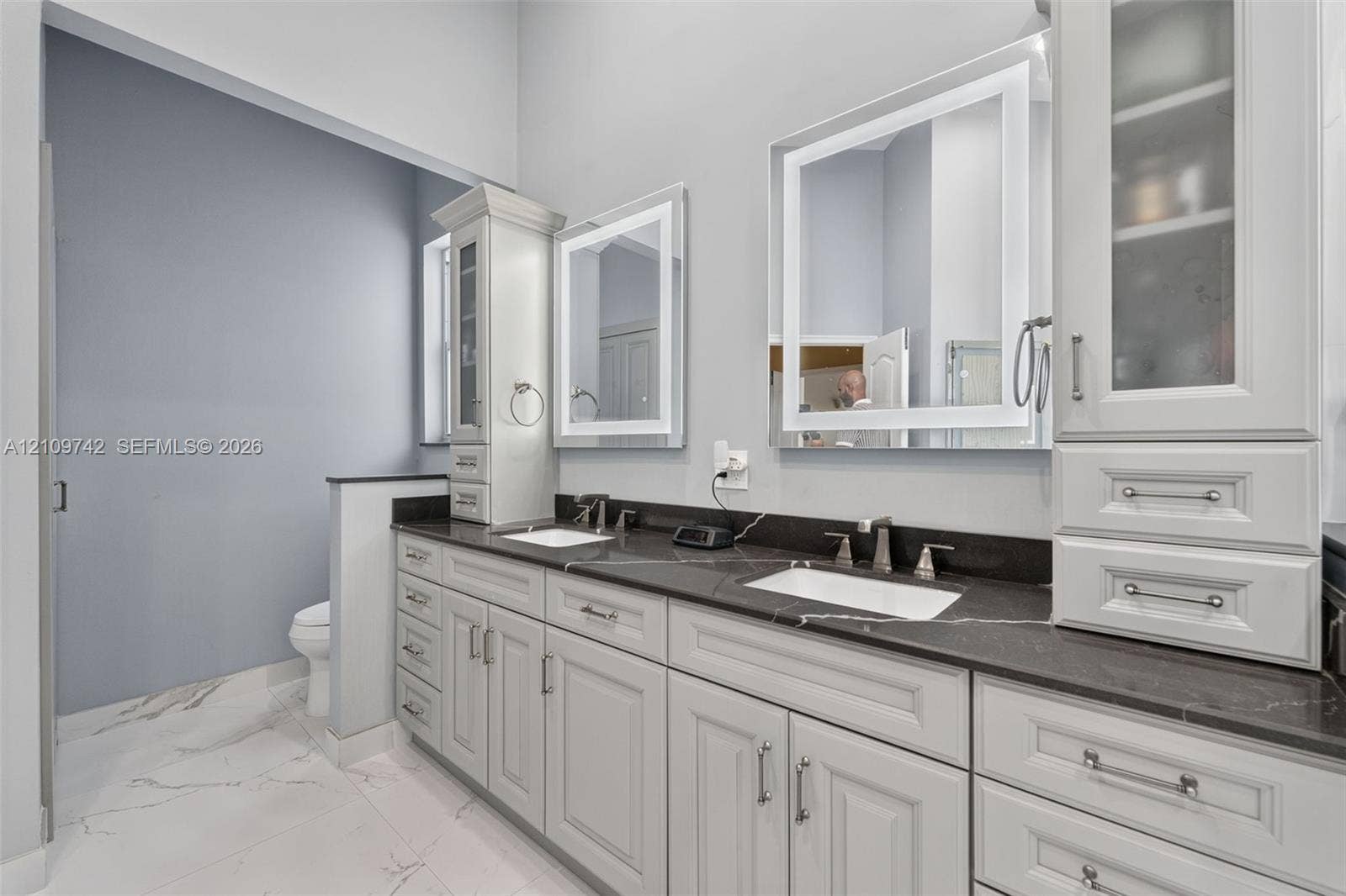 19143 SW 60th Court, Pembroke Pines, FL 33332 Dual-sink vanity with dark stone countertop, white shaker-style cabinetry, illuminated wall mirrors, and porcelain tile flooring
