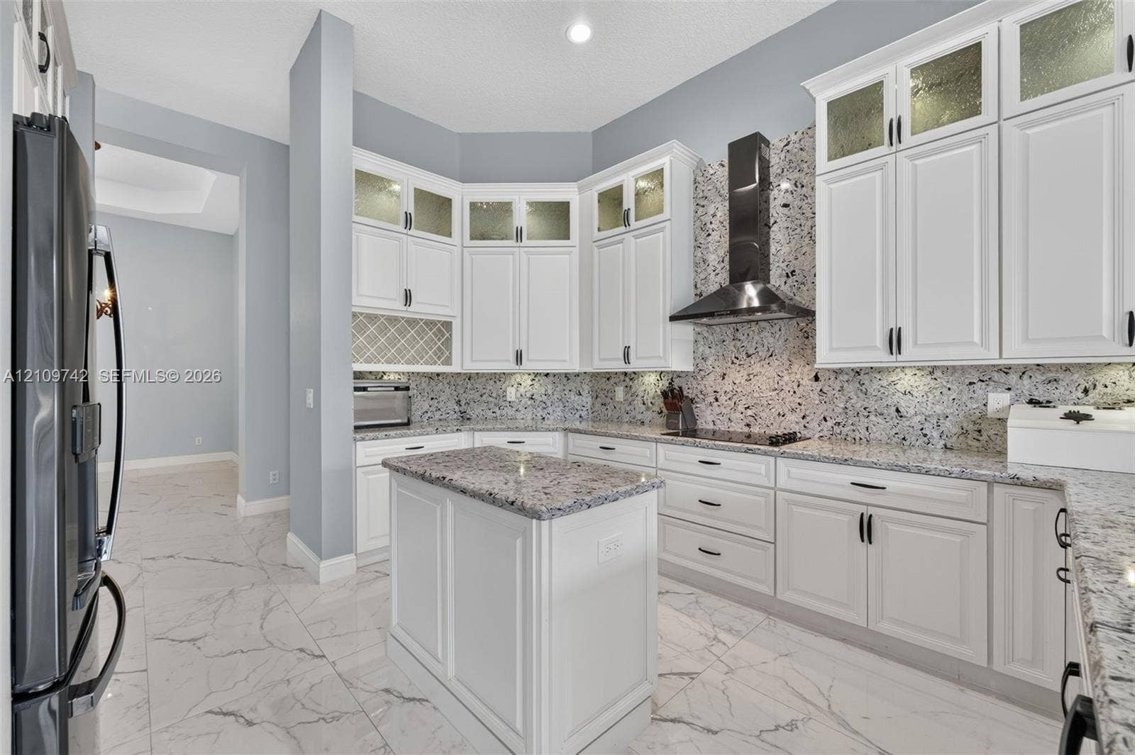 19143 SW 60th Court, Pembroke Pines, FL 33332 Kitchen featuring white shaker-style cabinetry with glass-panel uppers, patterned stone backsplash, center island, integrated cooktop with stainless steel range hood, and tile flooring