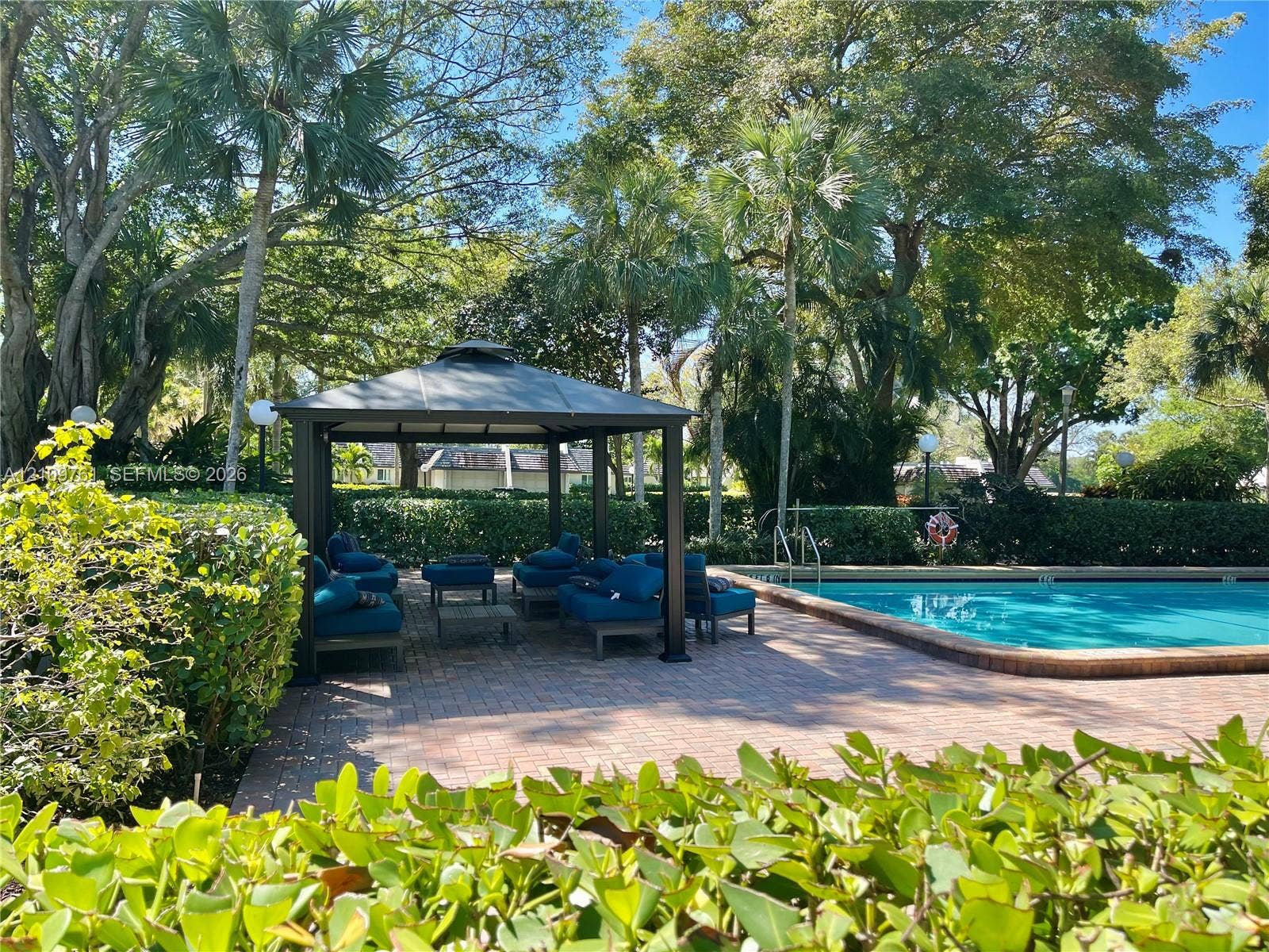 Outdoor swimming pool featuring a brick paver deck, permanent metal-frame gazebo, and mature landscape hedging