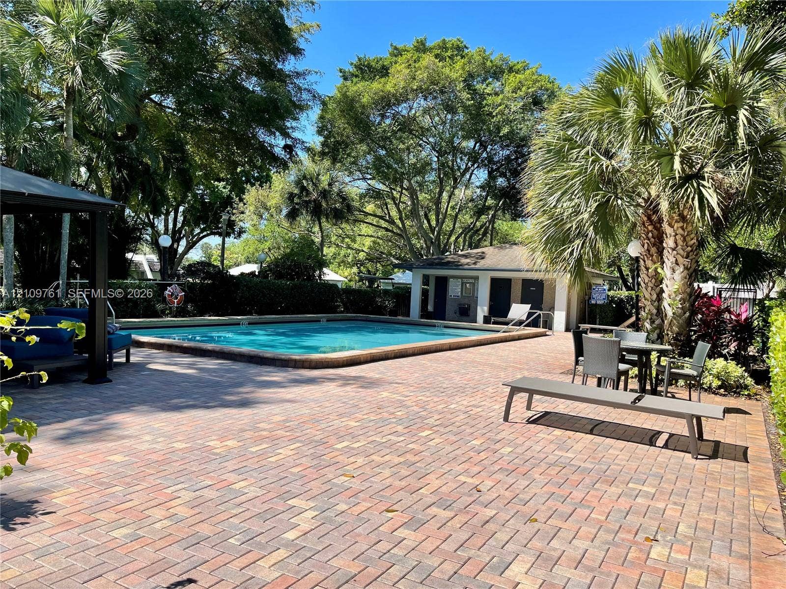 In-ground swimming pool featuring a brick paver deck, covered pavilion, mature palm trees, and established landscaping