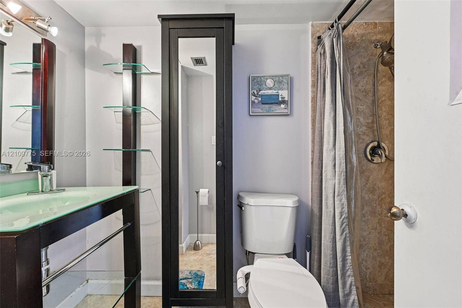 Bathroom featuring a glass-topped vanity with an integrated basin, a tall dark-finish built-in storage cabinet with a mirrored door, glass shelving, and a tiled shower stall with a hand-held shower head
