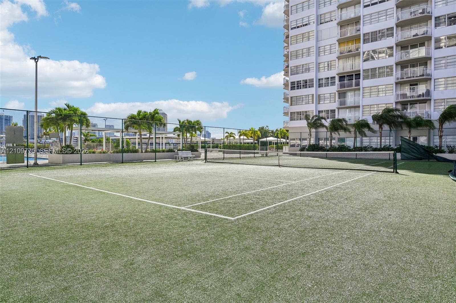 Tennis court with synthetic turf surface, enclosed by chain-link fencing, featuring overhead lighting and perimeter landscaping with palm trees