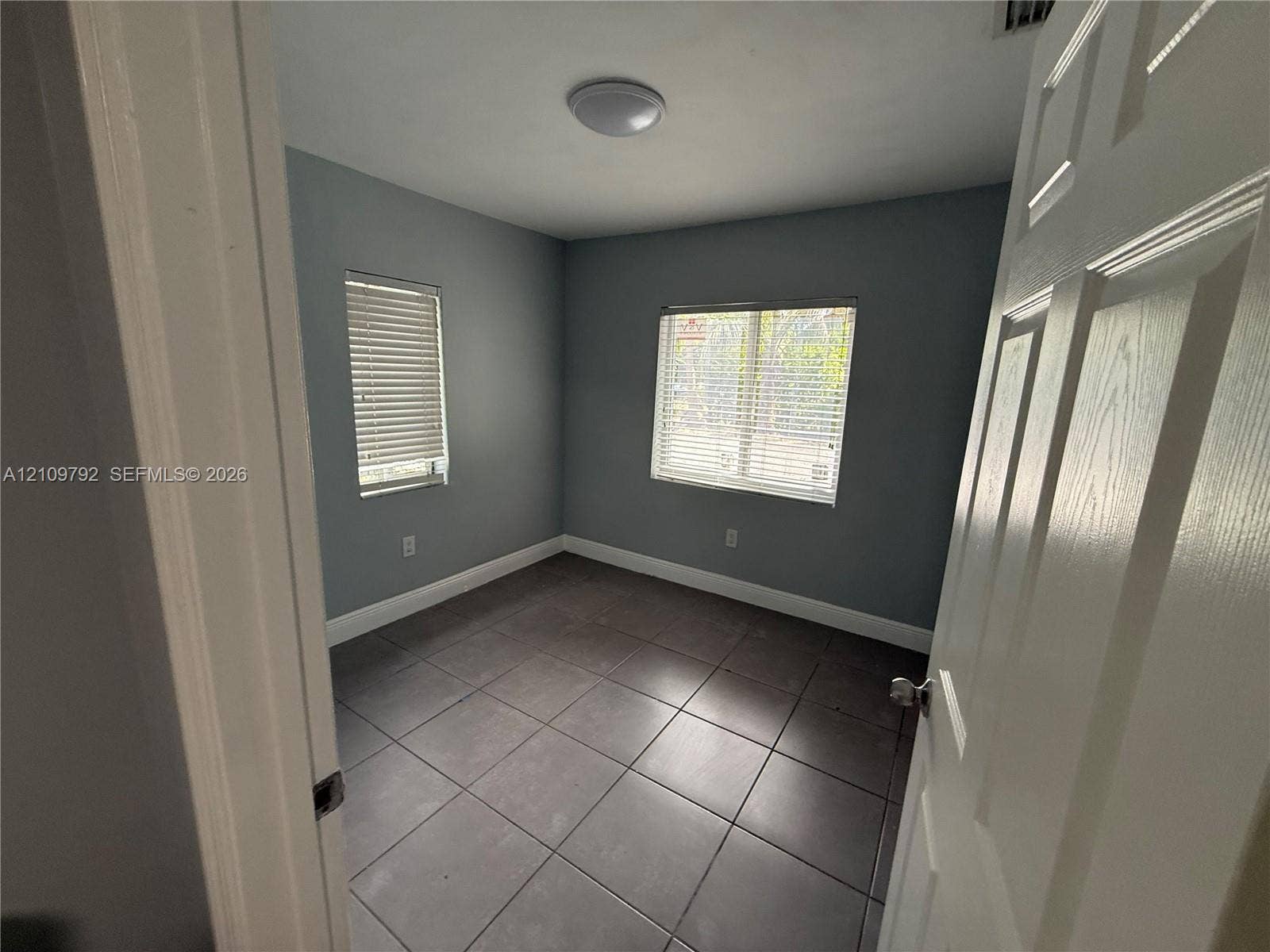 Room featuring large-format ceramic tile flooring, neutral grey wall paint, and two windows equipped with horizontal blinds