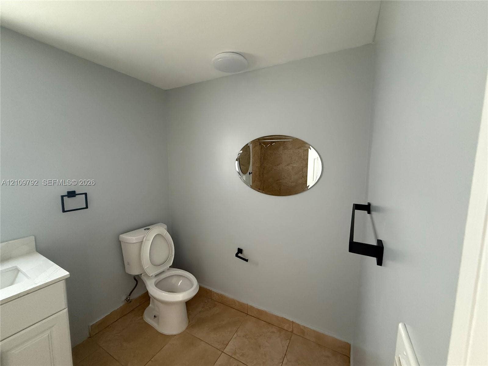 White vanity with stone countertop, oval wall-mounted mirror, matte black fixtures, and tiled flooring