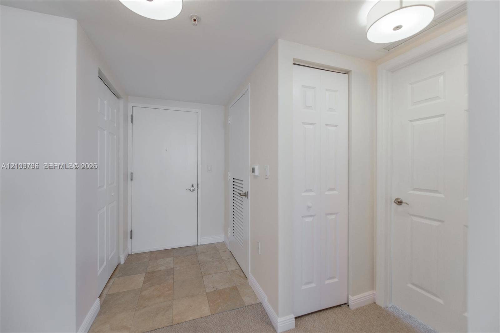 Entryway featuring stone-finish tile flooring, multiple multi-panel interior doors, a louvered mechanical closet door, and dual flush-mount circular lighting fixtures