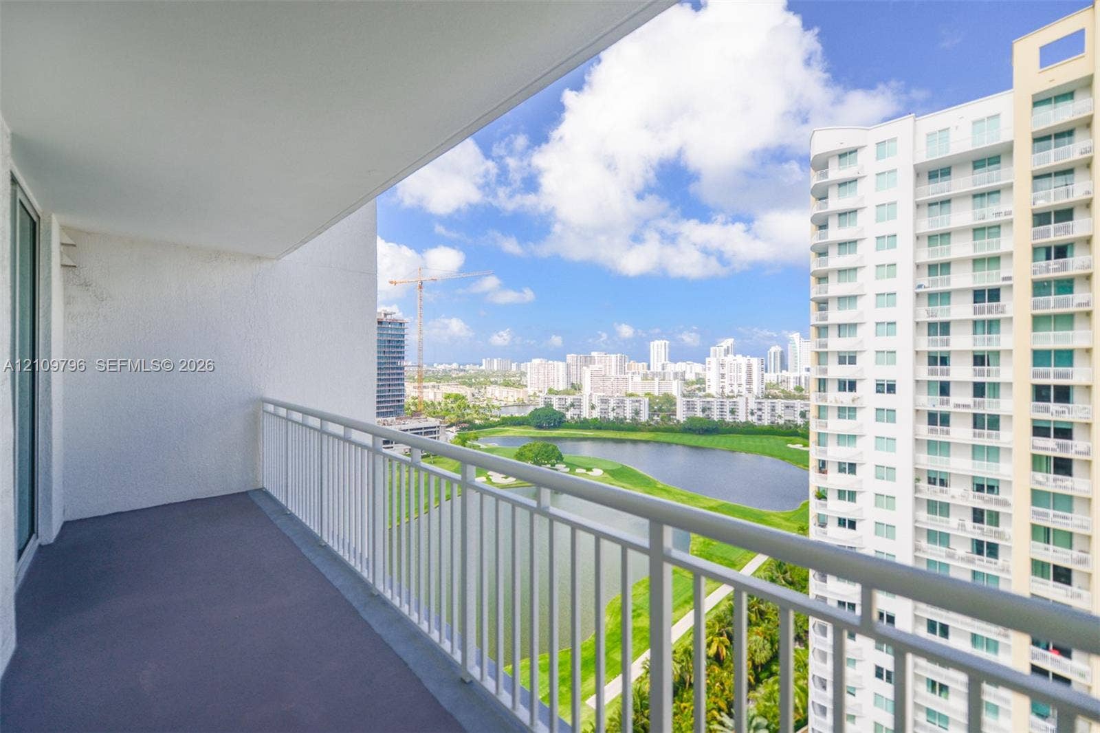 Covered balcony featuring a neutral-toned floor surface and white metal railings overlooking a lake, green space, and urban skyline