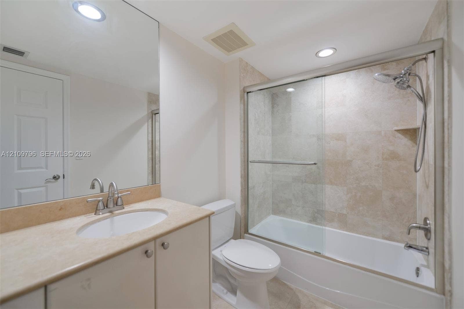 Bathroom featuring a vanity with a stone-finish countertop, undermount sink, full-width mirror, and a shower-tub combination with glass sliding doors and stone-tile wall surround