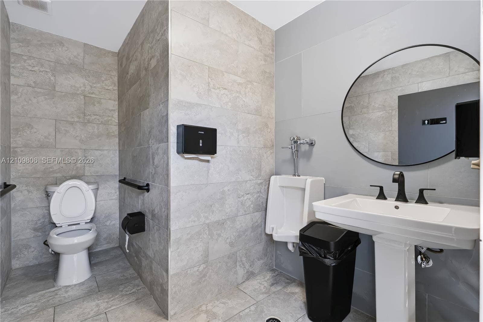 9123 SW 117th Court, Miami, FL 33186 Restroom featuring large-format neutral-toned wall and floor tile, wall-mounted porcelain urinal with manual flushometer, pedestal sink with matte black widespread faucet, and circular frameless mirror