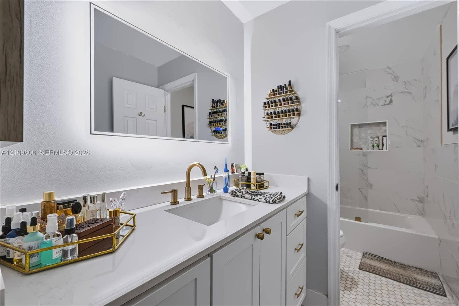 9123 SW 117th Court, Miami, FL 33186 Bathroom featuring a vanity with quartz countertop, undermount sink, and brass-tone faucet hardware, complemented by an integrated backlit mirror and a tiled shower surround with a built-in recessed niche