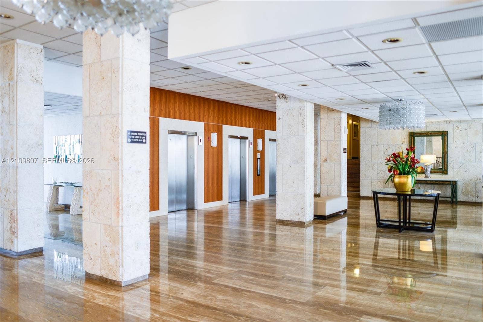 1980 S Ocean Drive, #16Q, Hallandale Beach, FL 33009 Lobby featuring polished stone flooring, stone-clad structural columns, wood-paneled elevator surround, suspended crystal-style light fixtures, and a recessed drop ceiling