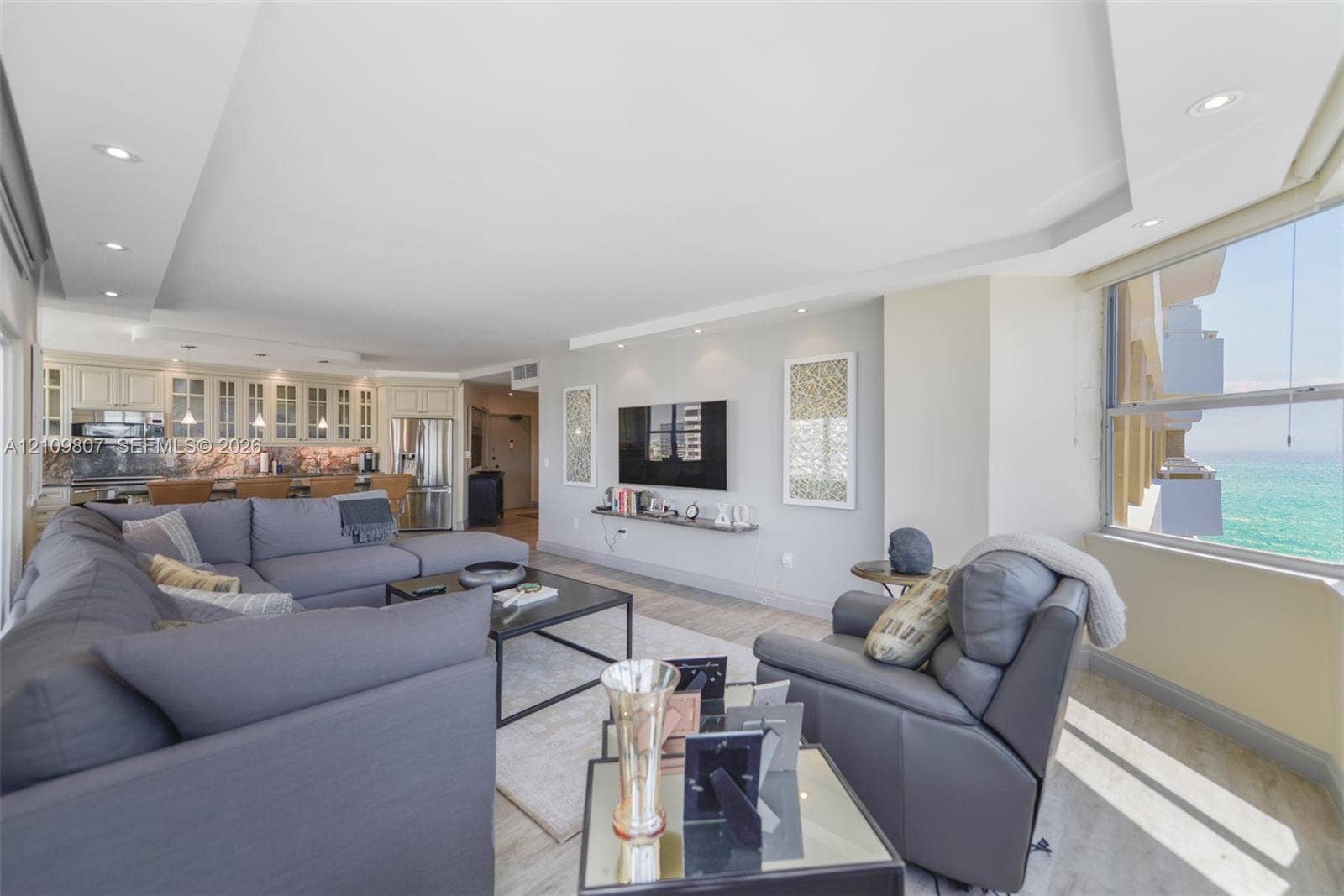 1980 S Ocean Drive, #16Q, Hallandale Beach, FL 33009 Open-concept living space featuring a tray ceiling with recessed lighting, wood-finish flooring, built-in kitchen cabinetry with glass-front uppers, and oversized windows